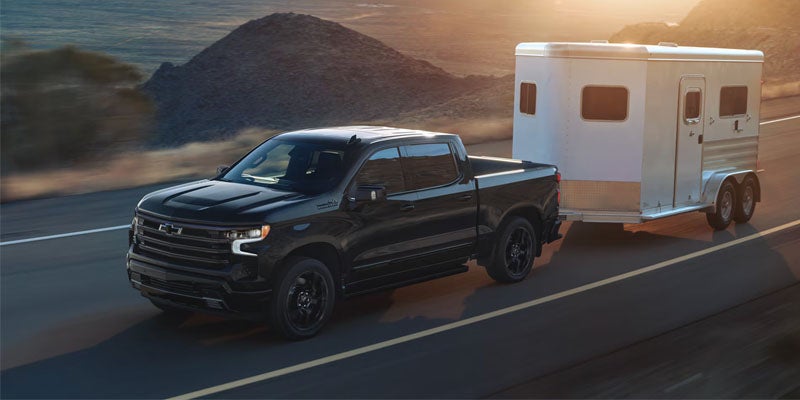 A black 2026 Chevy Silverado 1500 tows a white trailer behind as it drives down a highway at dusk