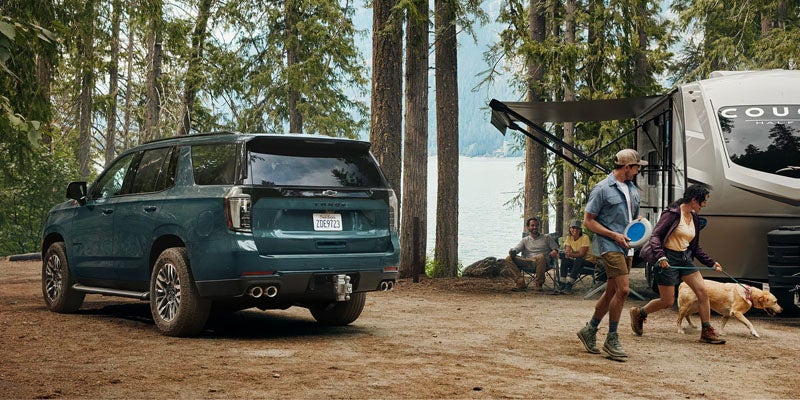 A seafoam colored 2026 Chevy Tahoe is parked at a campsite on a sunny day with woods, water, and a man and woman walking their dog near a camper is visible nearby the spacious SUV in Folsom, CA
